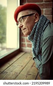 Old Age Concept. Portrait Of An Intelligent Elderly Man In A Beret, Scarf And Glasses Looking Thoughtfully Out The Window At Home. 