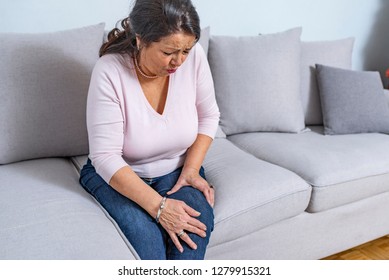 Old Age, Health Problem And People Concept - Senior Woman Suffering From Pain In Leg At Home. Senior Woman Holding The Knee With Pain. 