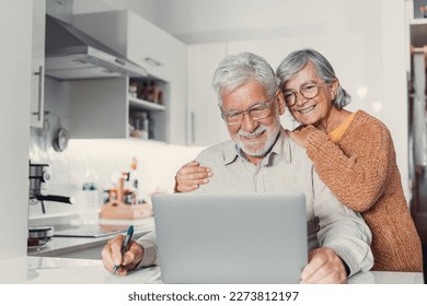 Old age and modern tech. Retired family couple sit at kitchen table use laptop watch photo video read latest news online. Aged husband share discuss interesting information on website with senior wife - Powered by Shutterstock