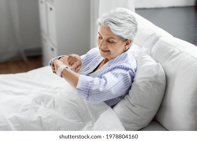 old age and people concept - happy smiling senior woman in pajamas with smart watch sitting in bed at home bedroom - Powered by Shutterstock