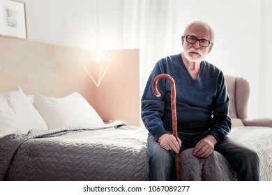 Old Age. Serious Nice Elderly Man Sitting On The Bed An Holding His Walking Stick While Resting At Home