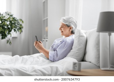 Old Age, Technology And People Concept - Happy Smiling Senior Woman In Pajamas Using Smartphone Sitting In Bed At Home Bedroom