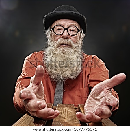 Old age. A very old man in old-fashioned hat and glasses with a long gray beard stretches out his hands asking for help. Studio portrait on a black background. 