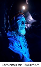 Old Alpinist With Grey Beard In Helmet With Glow Headlamp In Deep Dark Glacial Ice Cave With Blue Lights Explore Winter Mountain Glacier In Kazakhstan