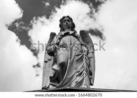 An old angel sculpture looking down in low angle shooting in top of mausoleum cemetery. Blue and cloudy sky at background. Black and White photography