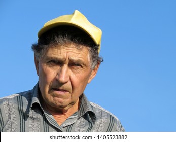 Old Angry Man Isolated On Blue Sky Background. Portrait Of Elderly Caucasian With Stern Face And Furrowed Brow, Villager In Baseball Cap, Concept Of Farmer, Retired, Disapproving Expression