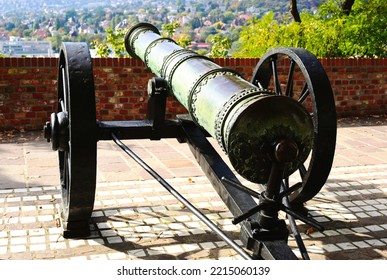 Old Antique Brass Cannon Closeup. Aged Patina Finish. Diminishing Perspective. Large Steel Wheel. Historic Fighting Equipment. Street Setting With Brick Fortress Wall. Decorative Pattern. Summer Scene