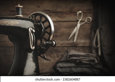 Old Antique Sewing Machine. In Background Is A Hanging Tailoring Scissors On Wooden Wall And Lying Cloths. Retro Tailor Workshop.