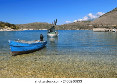 Old, artisanal, fiberglass fishing motor boats aground and moored in the shallow waters of Porto Palermo Bay on the Ionian Sea; Police speed boats docked at the background right pier. Himare-Albania.