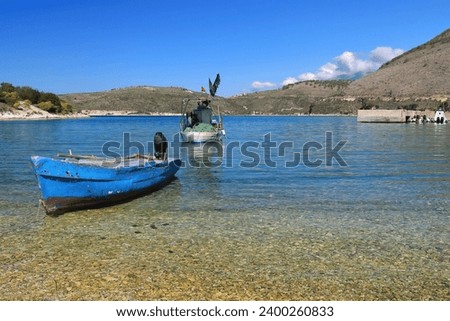 Old, artisanal, fiberglass fishing motor boats aground and moored in the shallow waters of Porto Palermo Bay on the Ionian Sea; Police speed boats docked at the background right pier. Himare-Albania.