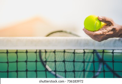 Old Asian Man Hold Two Tennis Balls In Left Hand, Selective Focus, Blurred Racket, Net And Green Tennis Court As Background, Aging Population Concept