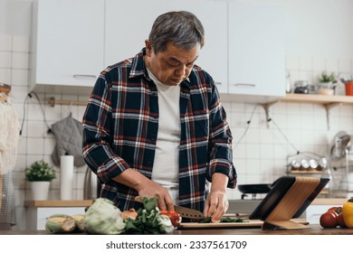 The old Asian man in the kitchen, using a digital tablet pad to learn cooking techniques, chopping vegetables on the board with ingredients in the home kitchen. - Powered by Shutterstock