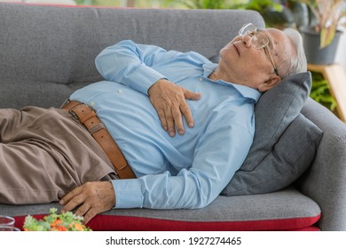 An Old Asian Man With Short Grey Hair Model Wearing Eyeglasses And Light Blue Long Sleeves Shirt Holding His Chest Around His Heart Lying Down On Sofa After Having Heart Attack Alone In His Home