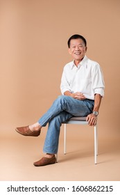 An Old Asian Man Sitting On Grey Chair And Smiling In Studio Beige Background.
