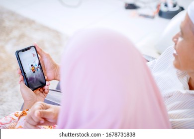 Old Asian Muslim Couple Holding A Phone, While Video Calling Their Grandchild In Their Home