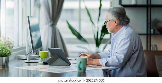 old asian senior retired age working at home hand ise desktop  and paper document at working desk in living room home interior background,asian male senior work at home