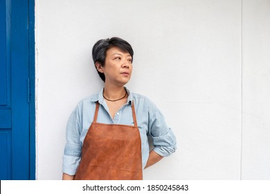 Old Asian Woman Wearing An Apron To Do Housework, Painting, Or Planing For Home Improvement Leaning Against The White Wall. Active Elderly Concept.