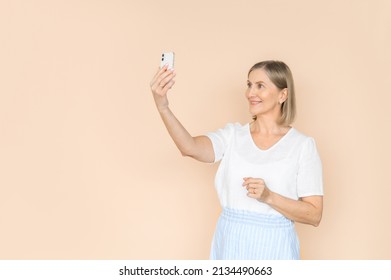 Old Attractive Woman Takes A Selfie On A Beige Background. Isolated Portrait In Light Clothes.