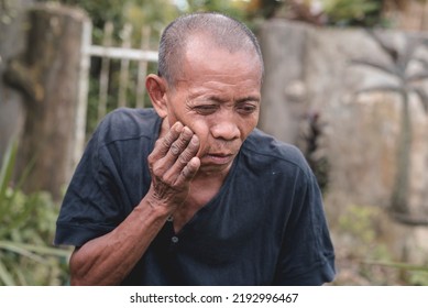 An Old Balding Asian Man Suffering From An Aching Molar. Attempting To Relieve The Pain. Experiencing A Bad Toothache Or Dry Socket While Outside.