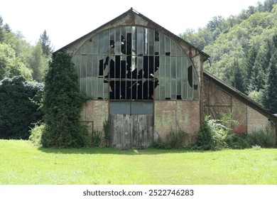 An old barn stands in a peaceful, green summer landscape, its wooden structure weathered by time.  Several windows are shattered, with broken glass clinging to the frames. - Powered by Shutterstock