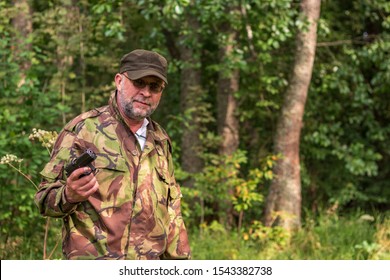 An Old Bearded Bandit With A Gun In His Hand. Russian Gangster In Camouflage Military Uniform. The Criminal Robs Walking In The Forest. A Man Is Engaged In Sports Shooting From A Traumatic Pistol.