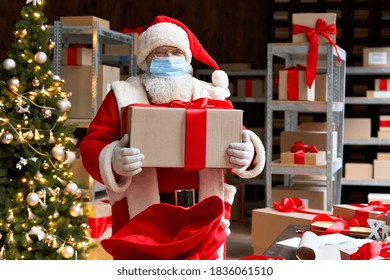 Old bearded Santa Claus wearing costume, face mask, holding present gift box packing in sack bag preparing for xmas eve in workshop. Merry Christmas Covid 19 coronavirus safe delivery concept. - Powered by Shutterstock