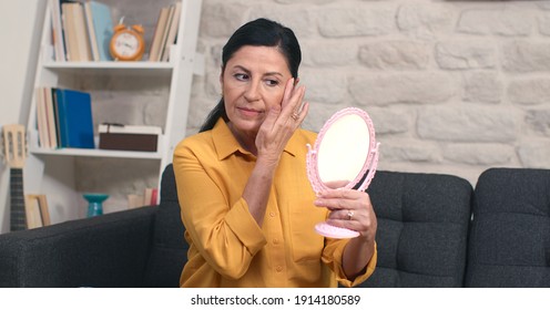 The old beautiful woman is looking at the small mirror in her hand, checking her skin and neck.