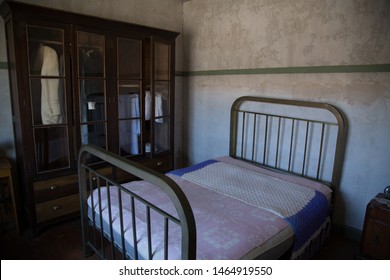 Old Bedroom, A Wardrobe With Glass Doors And Hanging Clothes Is Displayed, Also A Bed With Metal Frame