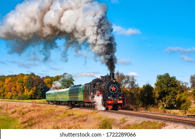 Old black steam train with green cars and much smoke. Vintage locomotive with historical tour. Railway in green and yellow autumn forest. top image from golden railroad times. Beautiful countryside