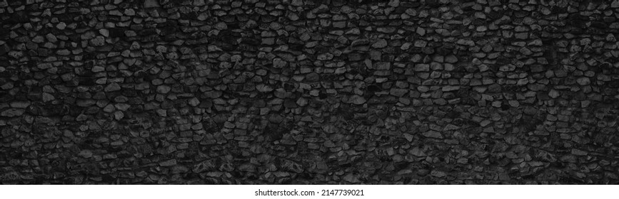 Old black stone wall wide panoramic texture. Rough crushed rock masonry gloomy backdrop. Dark gray abstract background