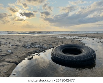 Old black tire lying in a shallow water pool on a rocky beach at sunset. Peaceful and moody seascape with soft light and dramatic clouds.