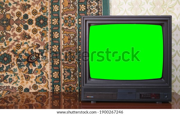 Old\
black vintage TV with green screen to add new images to the screen\
TV set against wallpaper and carpet from\
1980-1990.