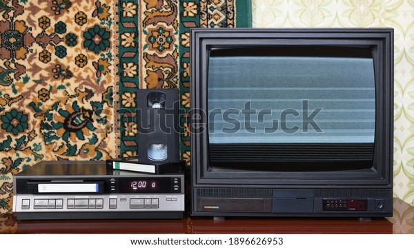 Old black\
vintage TV with noise and interference on the screen, VCR on the\
background of old carpet and\
wallpaper.