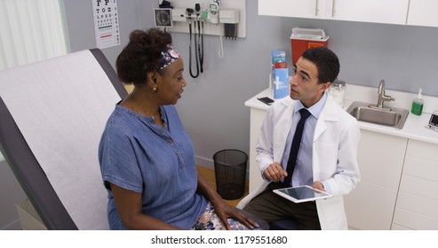 Old Black Woman Explaining Health Issue To Young Male Hispanic Doctor In Office