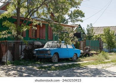 An old blue car with a trailer stands in front of a fence in the courtyard in the village under an acacia tree in the shade next to the path near the village house - Powered by Shutterstock