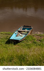 An old boat is in the grass by the river. There is sun, the water is brown.