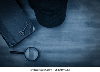 Old Book, A Key, A Detective Cap And A Magnifying Glass Against A Worn Wooden Background With Dark Edges Toned In The Classic Blue Color (top View) As The Crime Story Concept