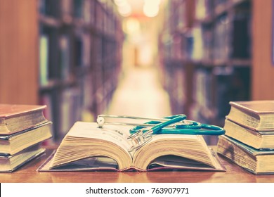 Old Book In Library With Stethoscope On Open Textbook, Stack Piles Of Texts On Reading Desk, And Aisle Of Bookshelves In  Study Class Room Background For Medical School Education Learning Concept