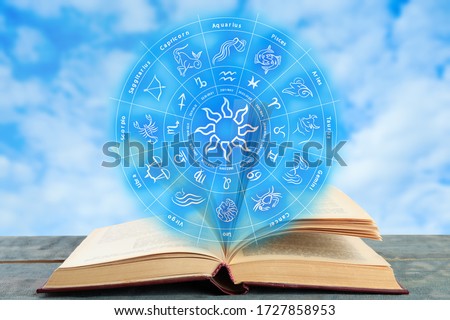 Old book on wooden table and illustration of zodiac wheel with astrological signs against blue sky