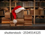 Old books and Santa Claus hat. Christmas readings concept. New Year in the library with stacks of books, bookshelves in the background. Literature for winter evenings during the holidays.