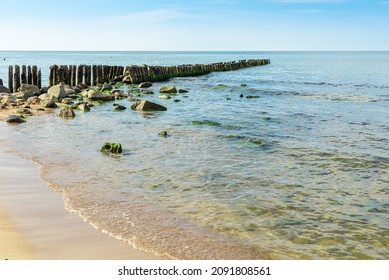 An Old Breakwater On The Sea. The Coastline With A Barrier For Waves. Breaking Waves On Wooden Poles. The Logs Of The Breakwater Covered With Mold. Stones Lying On The Coast. Seascape.