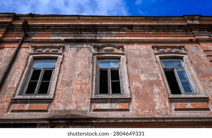 Old brick building facade with three windows. Weathered walls show peeling paint and decay. Blue sky visible above the roofline. Windows have broken panes and missing glass
