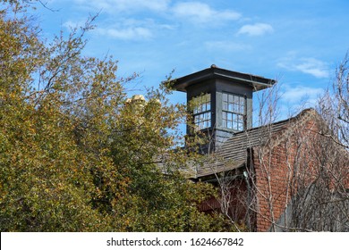 An Old Brick Building With A Glass Cupola And A Slate Tile Roof. Several Terracotta Chimney Pipes Are Visible Through The Tree Tops.