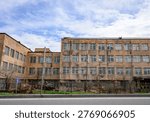 Old brick building under cloudy sky. Windows show signs of wear and neglect. Empty street suggests abandoned or underused facility. Ideal for urban decay, architectural study, or historical projects