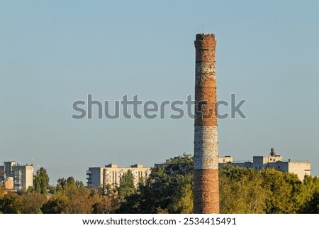 Old brick chimney on the background of the city skyline. A tall, weathered brick pipe stands prominently against the background of city buildings and greenery.