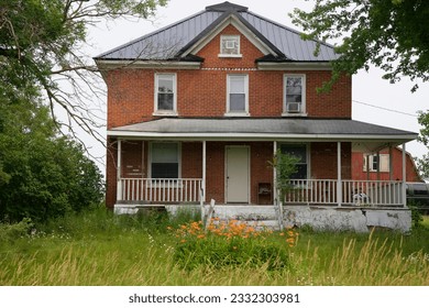 an old brick farm house building with a porch and a barn in the background, and flowers and tall grasses in the foreground - Shutterstock ID 2332303981