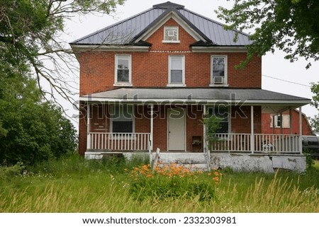 an old brick farm house building with a porch and a barn in the background, and flowers and tall grasses in the foreground