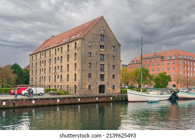 
An Old Brick House With Red Tiles And An LGBT Flag Hanging In The Window Near A Canal With Boat In The Neighbourhood Christianshavn. Copenhagen, Denmark