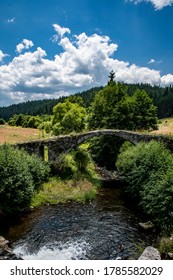 An old bridge in the middle of fields and a mountain with trees in the background. Blue sky with white clouds. High quality photo