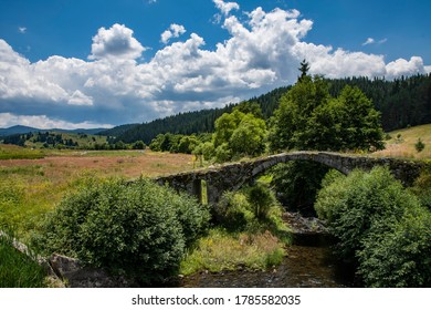 An old bridge in the middle of fields and a mountain with trees in the background. Blue sky with white clouds. High quality photo